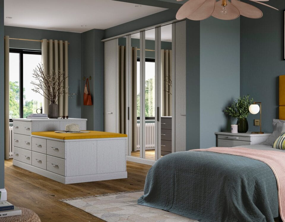A grey drawer island with a mustard-yellow top stands before mirrored sliding wardrobe doors in a dark teal Welford bedroom.