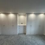 Bright fitted wardrobes line both sides of a carpeted bedroom, with white panelled doors, drawers and recessed lighting.
