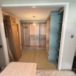 Fitted bedroom with warm wood wardrobes, open shelving, hanging rails and a pale carpet viewed through a doorway