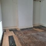 Unfinished bedroom with bare floorboards, patchy walls, a central chimney breast, skirting boards and a crystal ceiling light