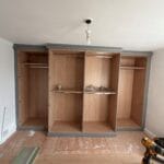 Unfinished fitted bedroom wardrobes with oak-effect interiors, grey trim, hanging rails and shelves beside renovation tools