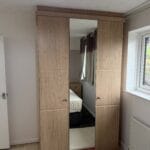 A wood-effect fitted wardrobe with two panelled doors and a central full-height mirror beside a bedroom window