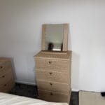 Oak-effect chest of drawers with four drawers and a framed mirror beside a bed in a neutral bedroom