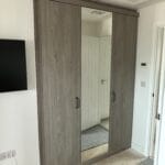A grey wood-effect fitted wardrobe with sliding doors and a central mirror stands beside a white bedroom doorway.