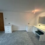 A bright white fitted bedroom includes a double dressing table, drawers, wall mirror, grey carpet and a wooden door.