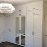 A white fitted bedroom with mirrored wardrobe doors, overhead cupboards, panelled doors and a sparkling crystal chandelier.