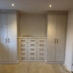 Light wood fitted wardrobes and a central chest of drawers create a balanced bedroom storage arrangement.