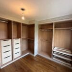 Bespoke fitted wardrobes with wood-effect interiors, cream drawers, hanging rails and open shelving around a bedroom corner