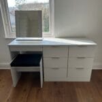 White dressing table with mirror, drawers and padded stool beside a sunlit window in a bedroom with wooden flooring.