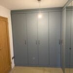 Custom grey fitted wardrobes span a bedroom wall beside a wooden door, with mirrored panels and a beige carpeted floor.